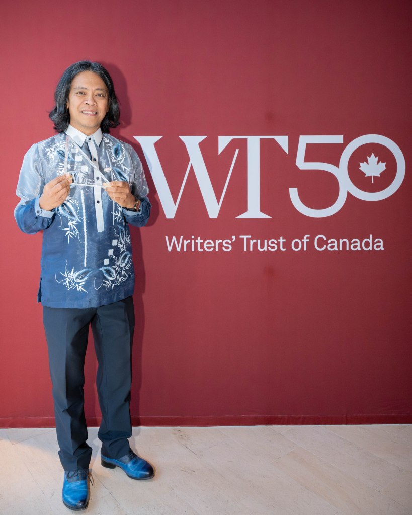 A person in a blue embroidered shirt stands in front of a red backdrop that reads 'WT50 Writers' Trust of Canada,' holding an award.